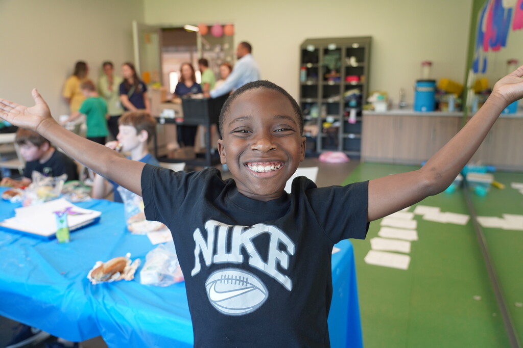 A boy with arms outstretched, smiling, wearing a black Nike shirt, at an event with tables and people.