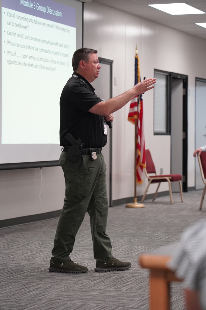 Man in green pants and a black shirt stands in a classroom and gestures toward a projector.