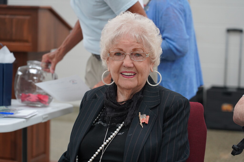 An elderly woman with white hair wearing glasses, a black coat, and pearl necklace smiles. She sits in a red chair at an event.