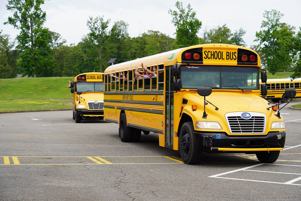 Two yellow school buses in a parking lot, one moving, with green trees and grass in the background.