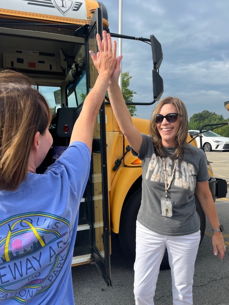 A happy woman in white pants and a gray shirt greets a teacher with a high-five in front of a yellow school bus.