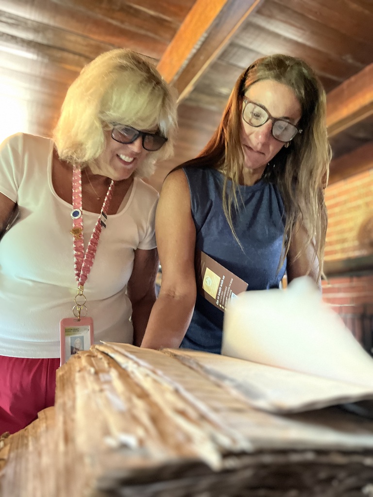 Two female teachers turn pages on a old book that features artifacts in a log cabin that serves as a museum. 