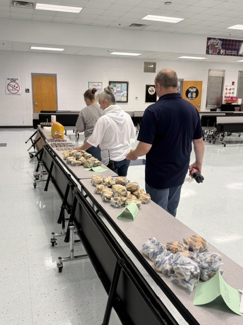A group of people are in a room with a long table displaying food. A man in a blue shirt is standing at the table.