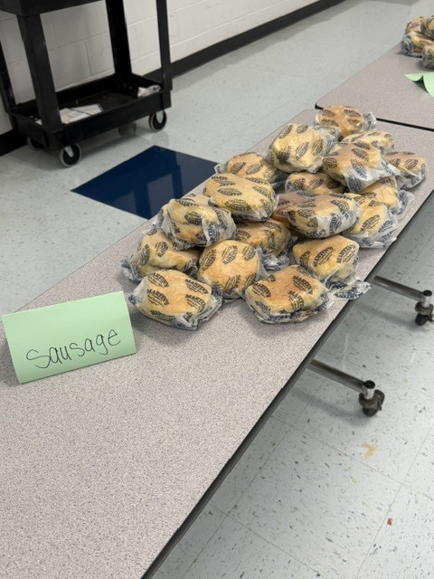 A table displays many individually wrapped items. A green sign with the word "Sausage" is on the left.