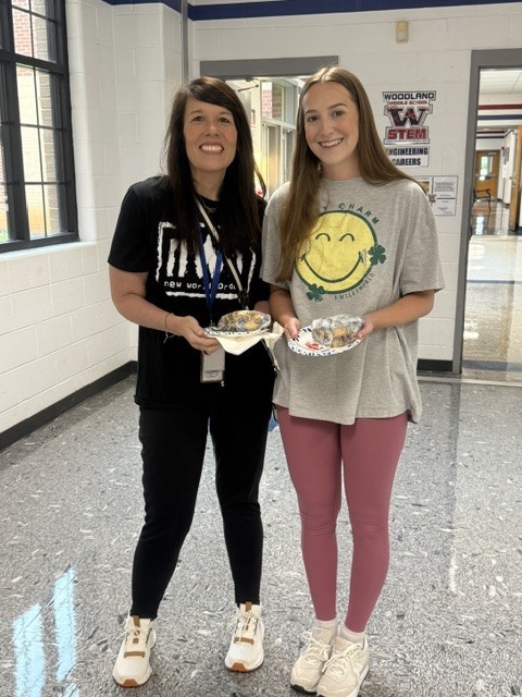 Two women stand in a hallway holding food, one in black, the other in gray with a smiley face.