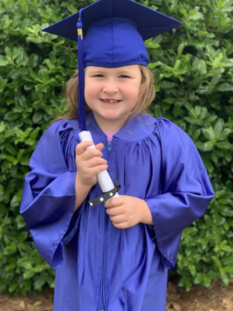 Child in blue graduation gown and cap holding a rolled diploma. Background of green shrubs.