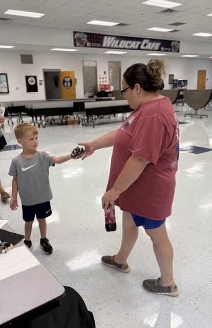 Woman and child stand in a cafeteria. The woman holds a red cup while the child holds a black item.