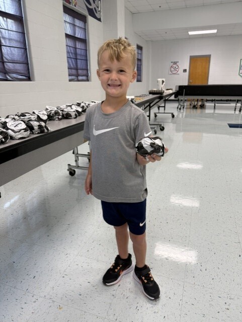 Child in sports gear stands in a room with white floor, holding a football. Long table with helmets nearby.
