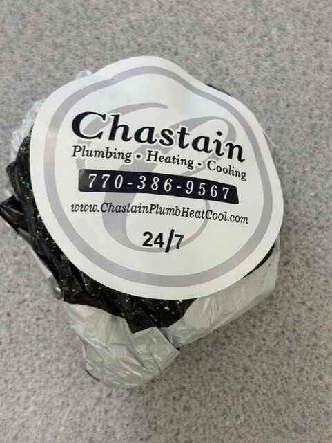 A cylindrical item wrapped in black and silver plastic. It has a label with white text reading "Chastain Plumbing Heating Cooling" and contact information.