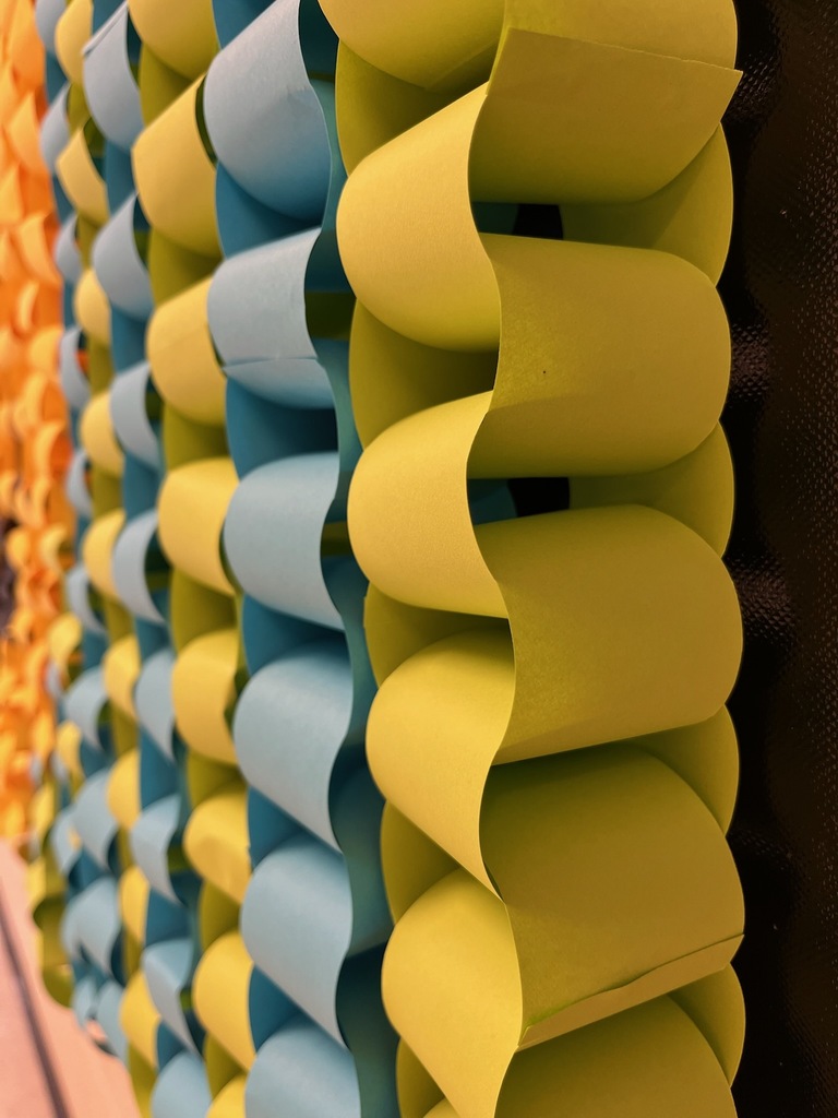 Close-up view of a bulletin board covered with interlocking paper chain links. The links are made from alternating strips of yellow-green and light blue paper arranged in vertical rows. The photograph is taken at an angle, highlighting the curved shapes, depth, and repeating pattern created by the paper loops. No text is visible in the image.
