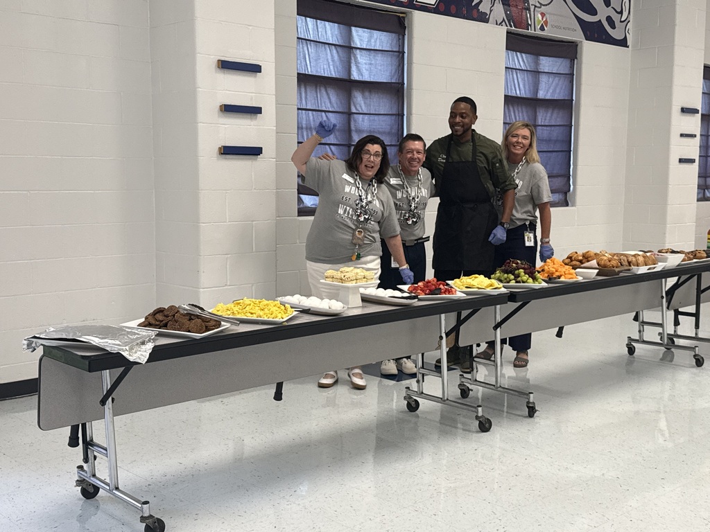 Four people stand behind a long table with various foods, wearing aprons and gloves. The room has white walls and large windows.