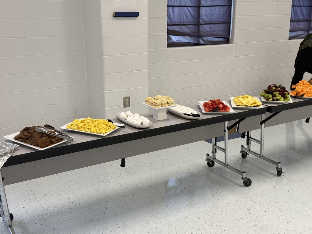 Long table with various foods on white plates, including desserts, fruits, and cookies. White walls and windows in background.