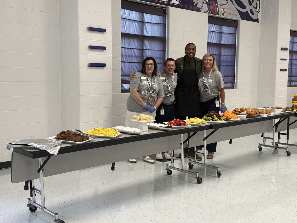 Four individuals stand behind a long table filled with various foods, including fruits, bread, and other dishes.