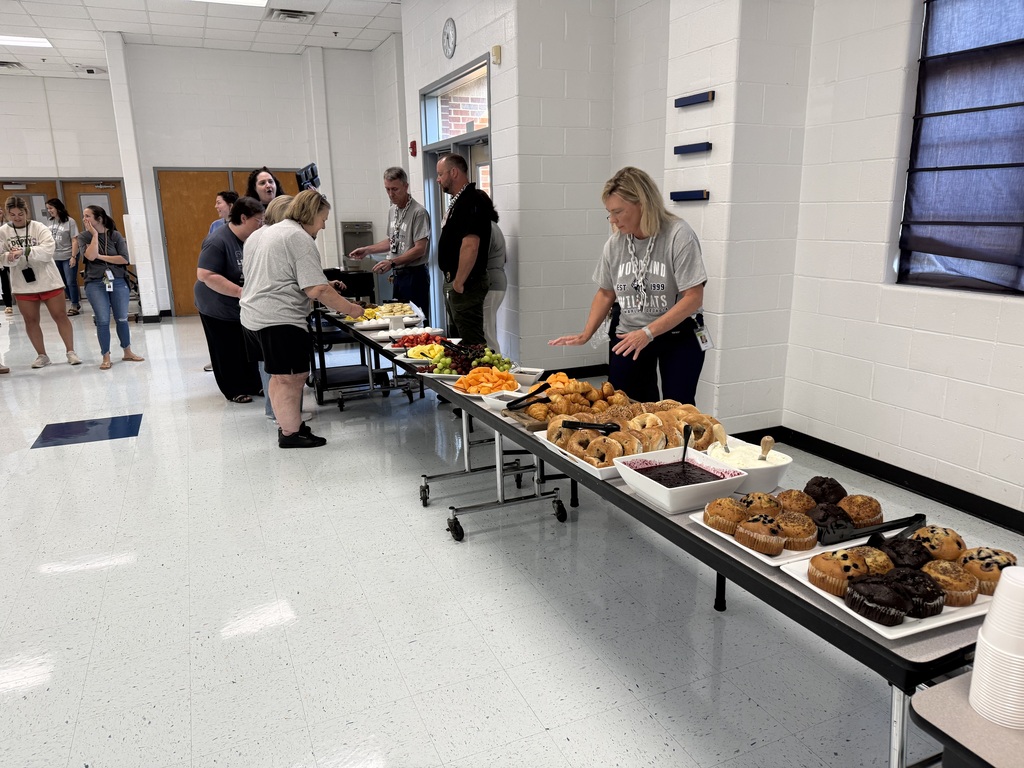 People are standing around a buffet table with trays of food and desserts, in a white-walled room.