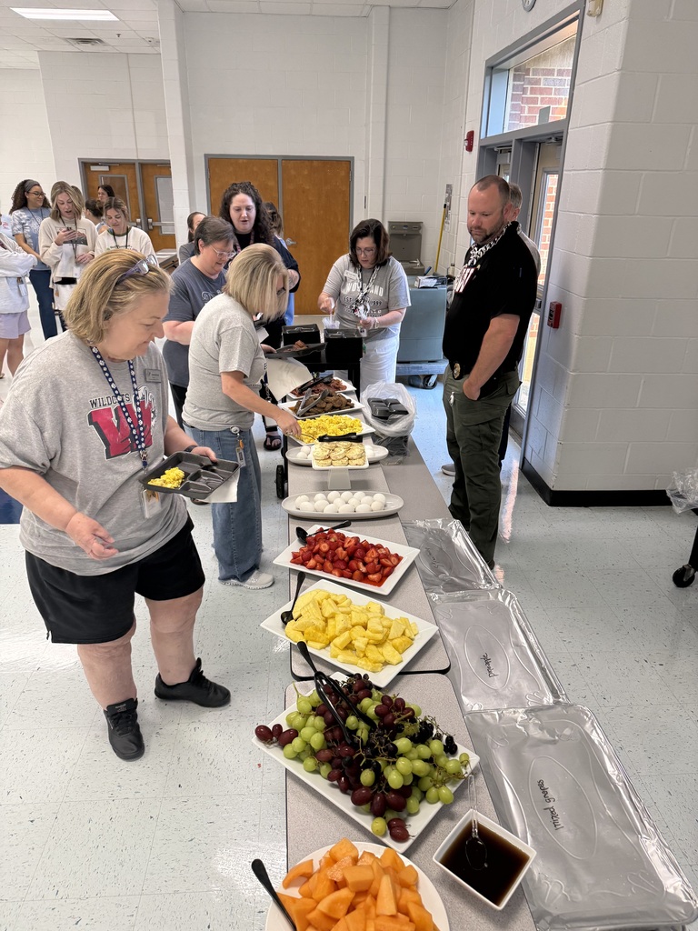 People stand at a buffet table with various fruits and foods in a room with a white floor.