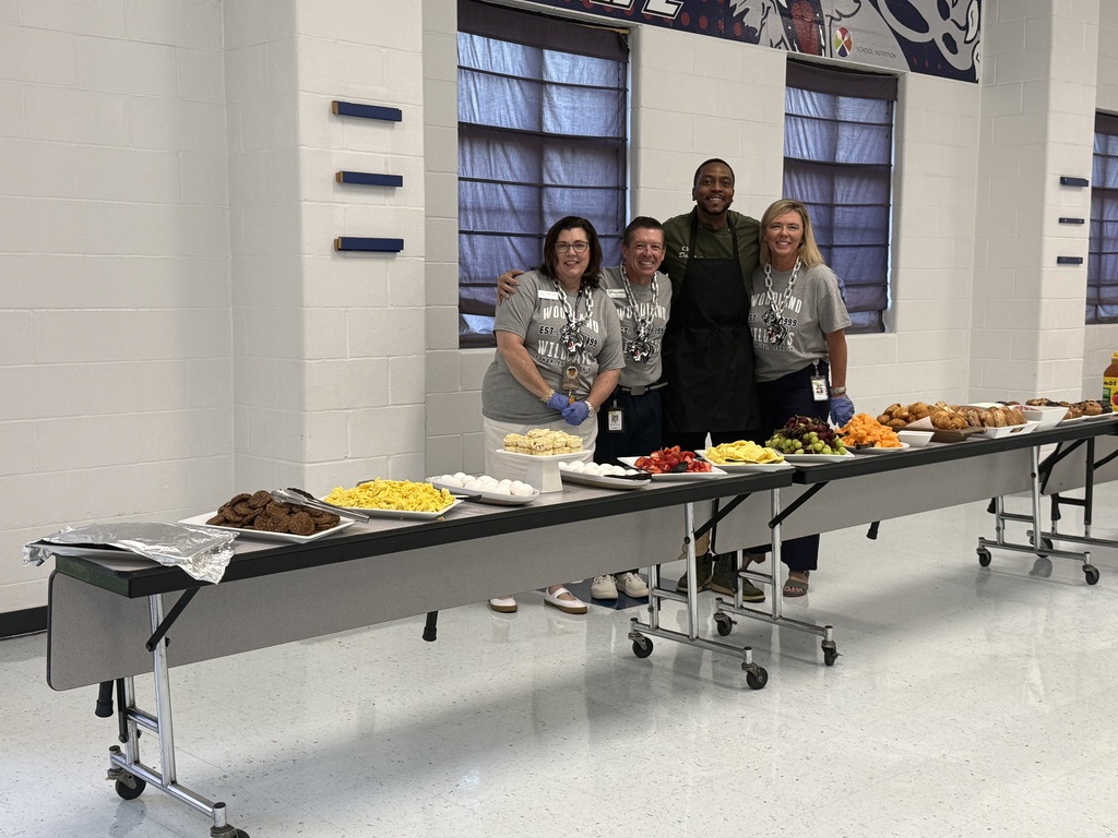 Four people in aprons stand behind a long table with various foods on it, such as fruit and sandwiches.