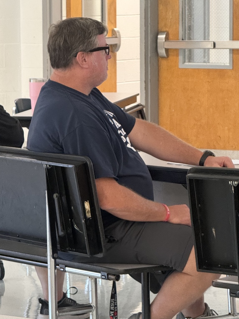 A man with glasses seated in a chair, wearing a blue shirt and black shorts. The room has wooden doors and white walls.