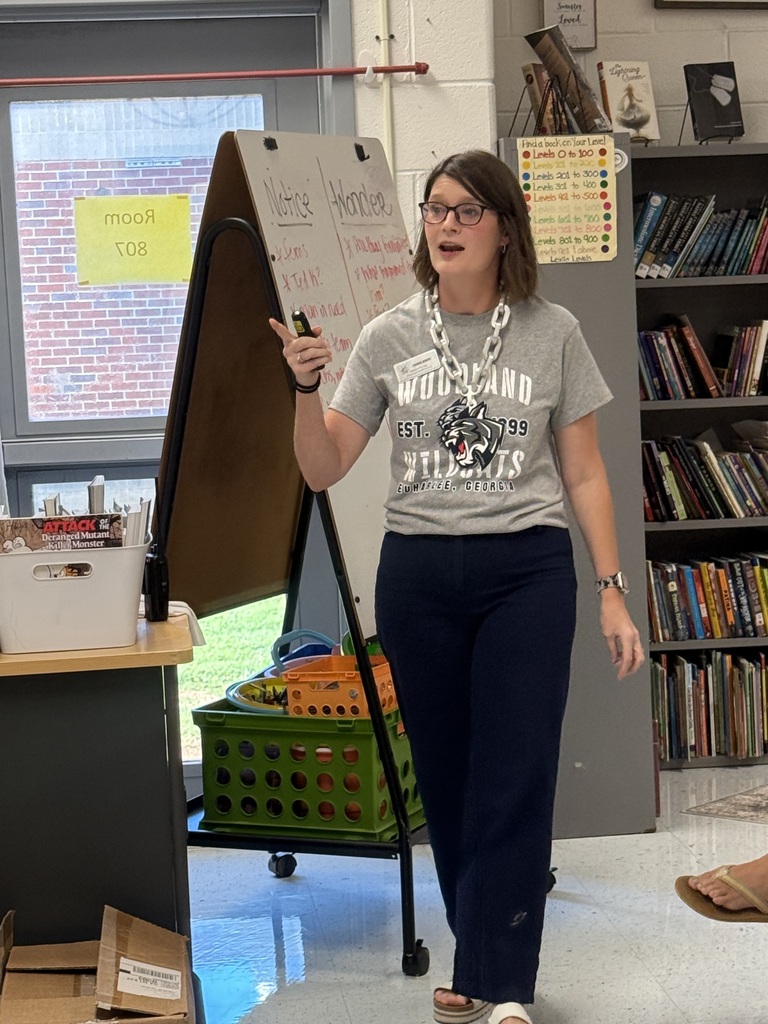 A woman in a gray t-shirt, glasses, and a necklace walks in a classroom, holding a remote. Behind her, a board stands on a stand near a desk.