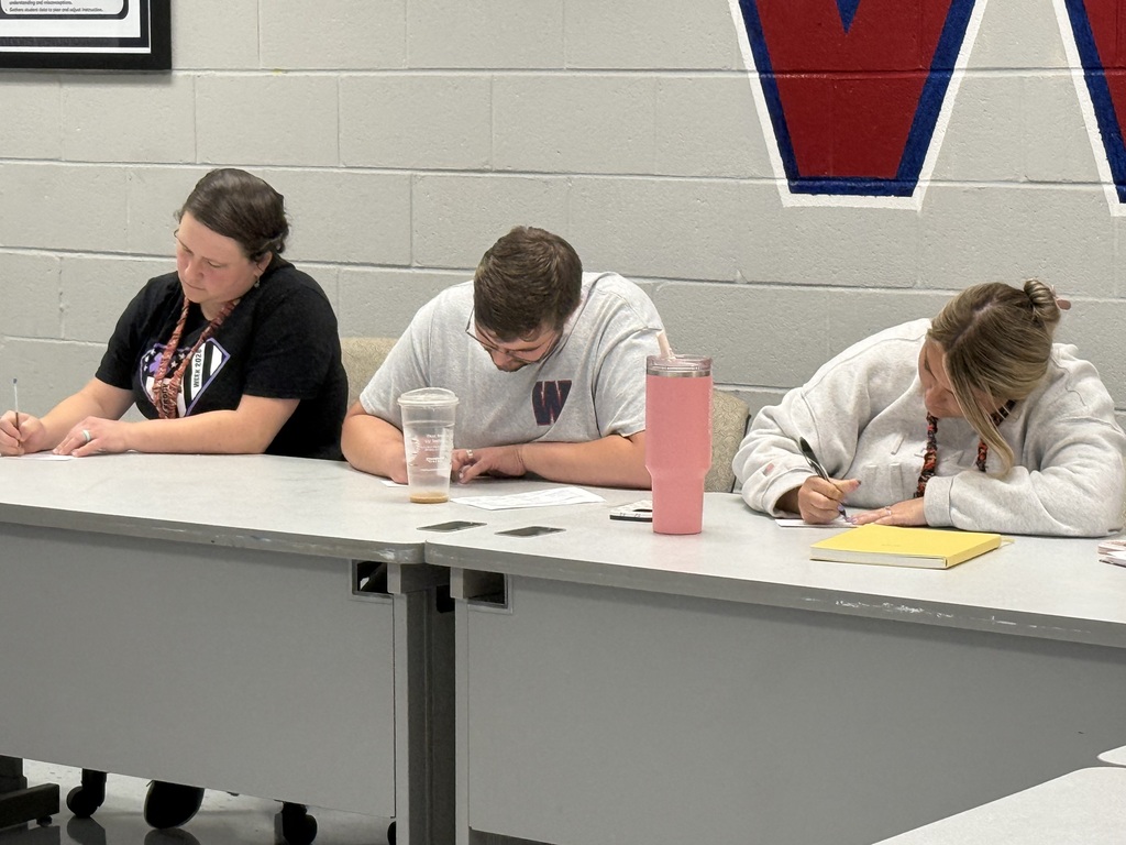 Three people sit at a table in a room with a white wall. Two wear glasses. They hold pens and write.