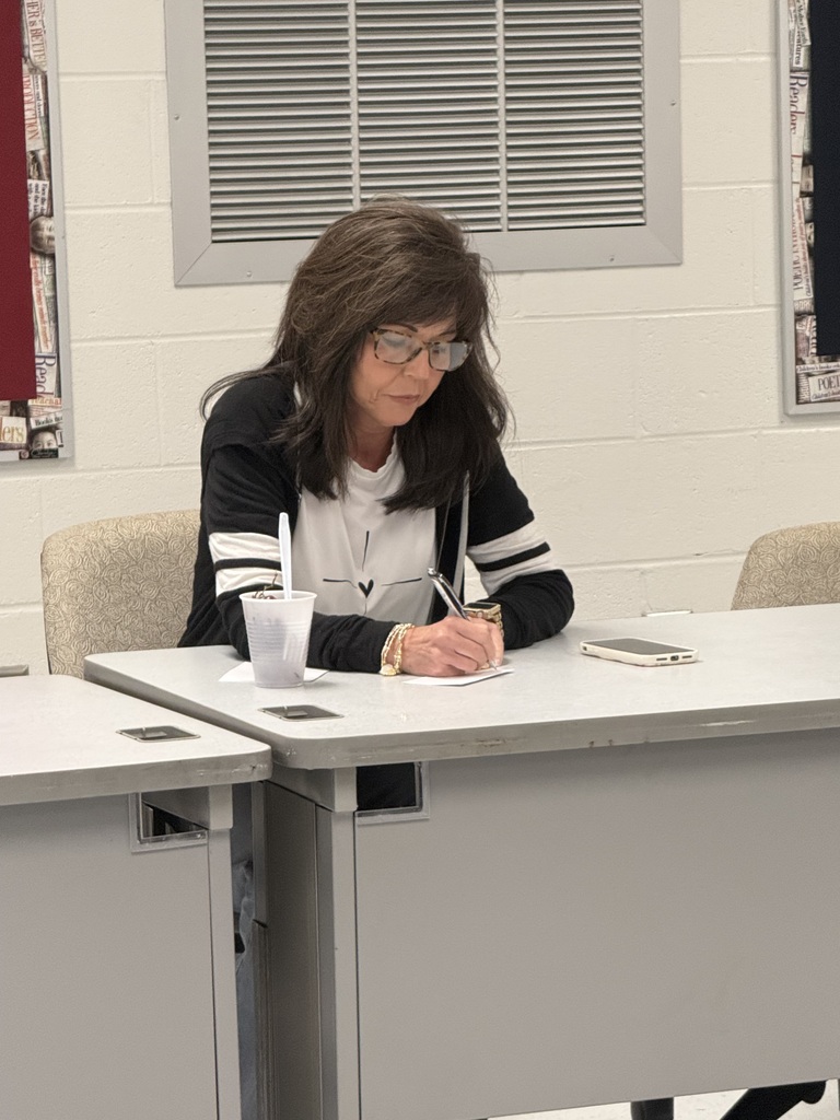 A person wearing glasses and a bracelet sits at a desk, writing with a pen.