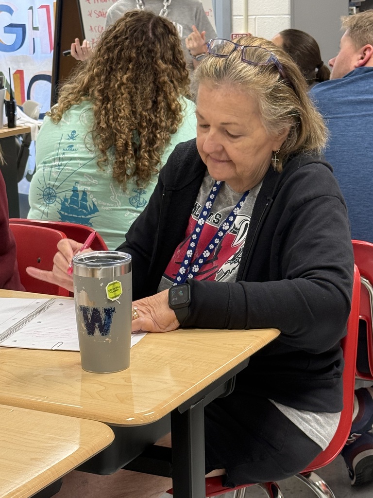 An older woman sits at a desk, writing with a pen. A gray tumbler with the letter W is in front of her.