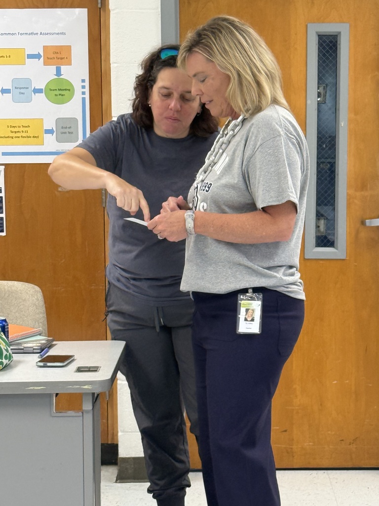 Two women in a room, one looking at a phone while the other points at a bulletin board.