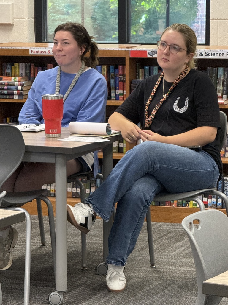 Two women seated in a classroom, one with a red cup on the table, the other with a necklace.