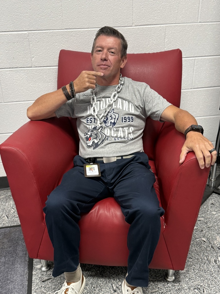 A man with a chain around his neck sits in a red chair. He wears a gray shirt and blue pants.