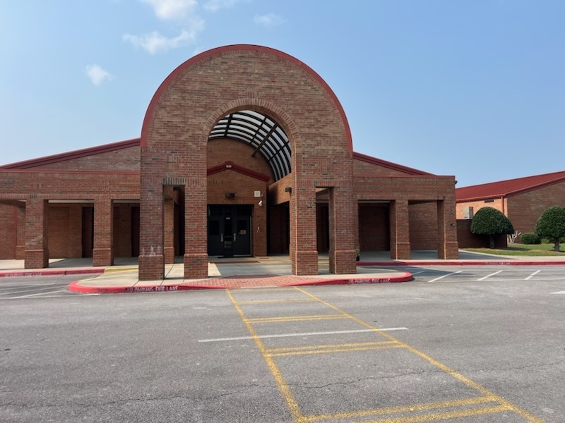 Front entrance of Pine Log Elementary School. The brick school building features a large arched entrance with a covered walkway leading to dark entrance doors. Red metal roofing is visible above the brick structure, and a parking lot with painted traffic markings is in the foreground. The sky is blue with a few clouds.