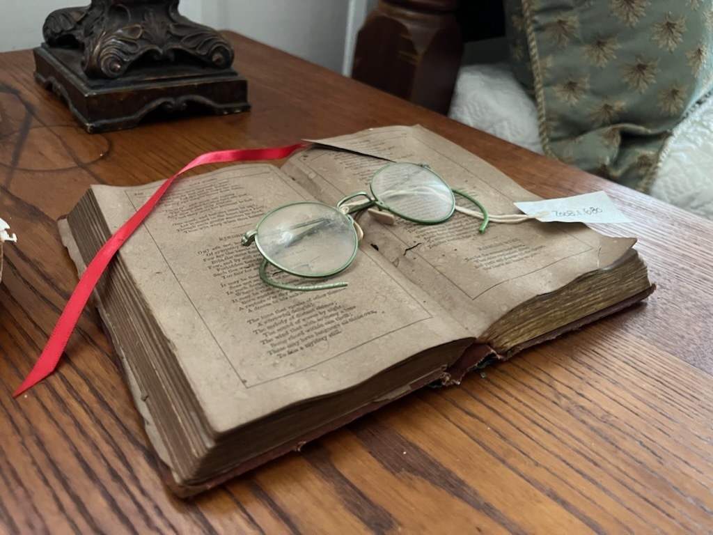 An open book with a pair of antique eyeglasses sits on a wooden table. A red ribbon is draped across the book.