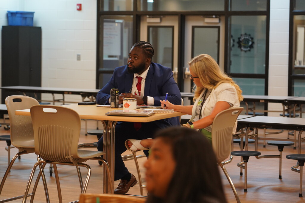 Two people in a school cafeteria, one seated at a table, another standing, both engaged in a discussion.
