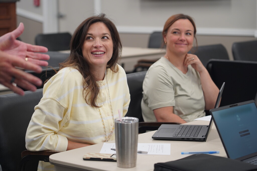 Two women seated at a table, smiling, with laptops and office items in front of them.