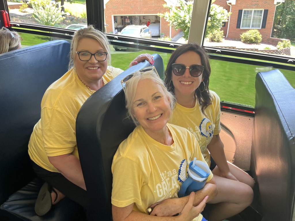 Three women in yellow shirts sit on a bus. They smile at the camera. Windows show a house and garden.