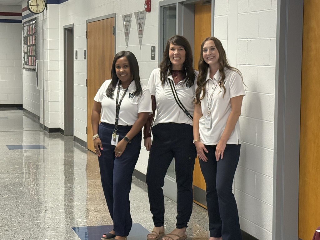 Three women in white shirts and blue pants stand in a hallway. A woman in the center has a scarf around her neck.