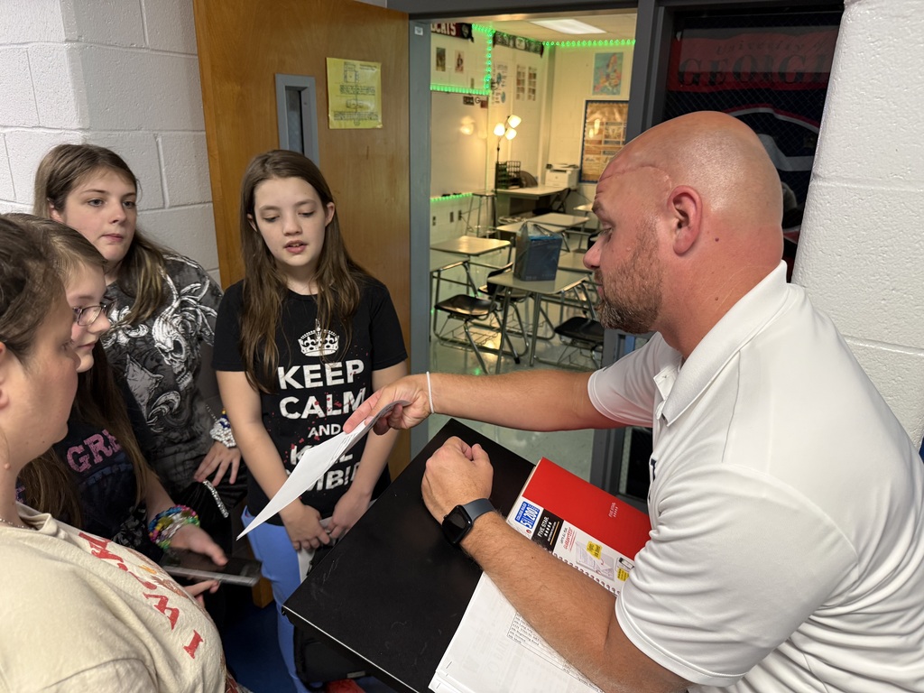 Man with bald head in white shirt hands paper to students in a classroom with tables and chairs.