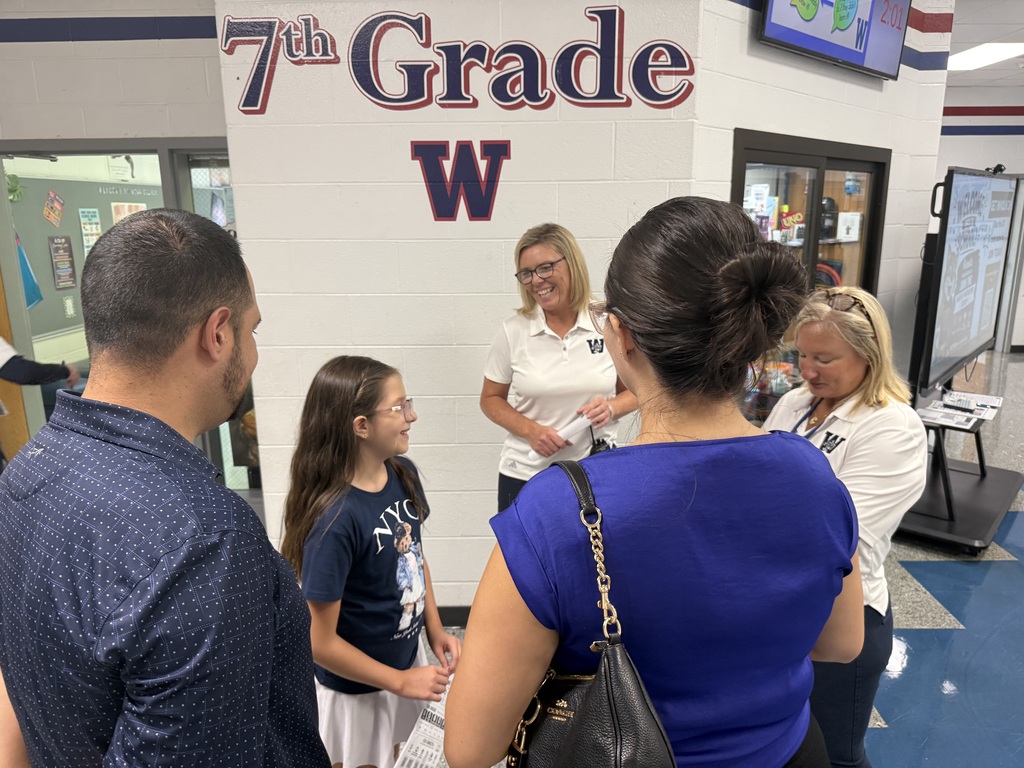 Five people stand in a school hallway. A woman in glasses speaks to a girl in front.