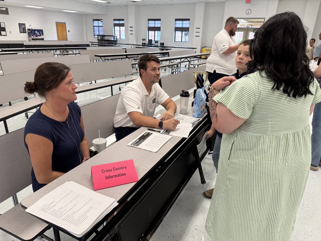Three adults and two children in a room with chairs. Two adults sit at a table, one standing.