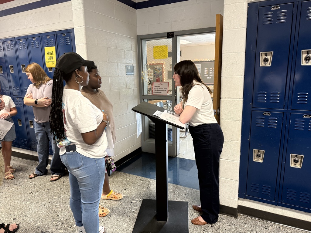 Group of people in a school hallway, one reading from a podium while others listen, lockers in background.