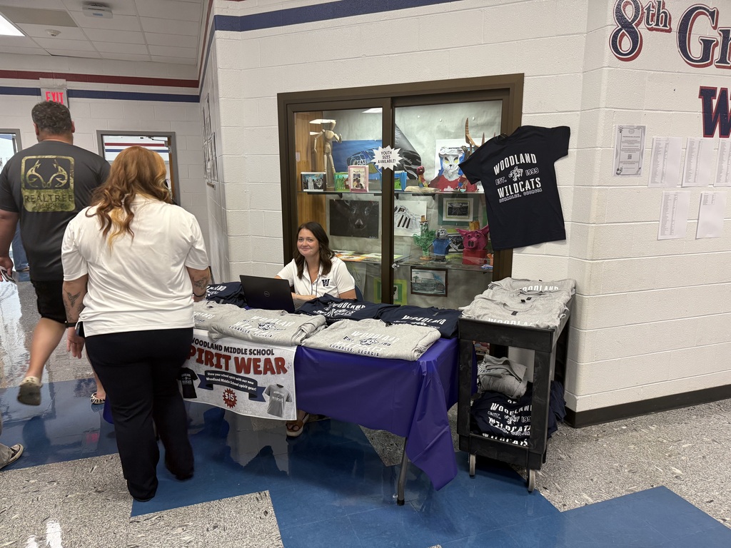 A woman sits at a desk with a laptop, while two people walk by. The table displays a sign for "Terry Wear" and a blue cloth.