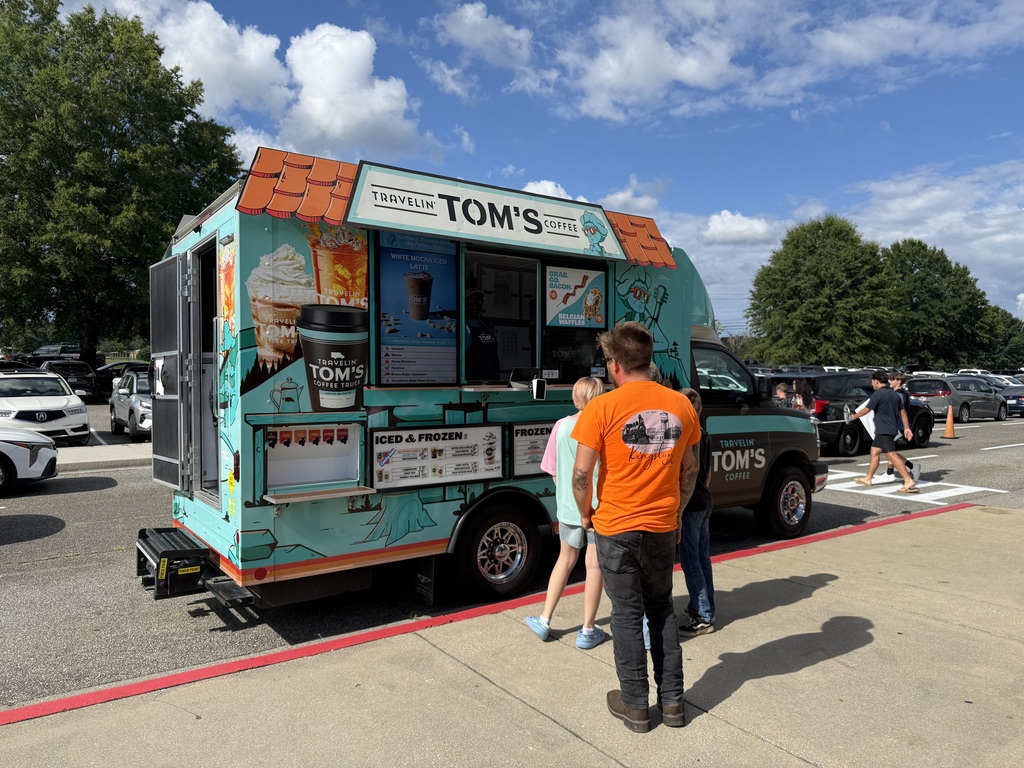 A food truck with "TOM'S" written on it is parked in a parking lot with trees. Three people are standing by it.