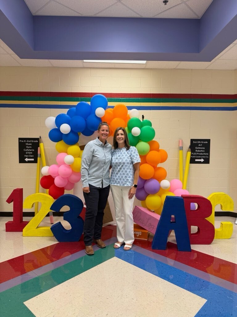 Two smiling women wearing blue tops and pants stand in front of a colorful balloon arch and letters spelling out 123 ABC.