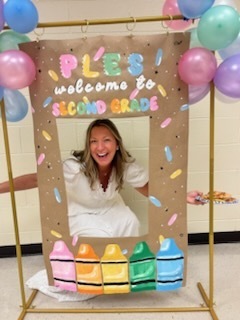 A smiling woman in a white dress crouches behind a photo booth backdrop reading "PLES welcome to SECOND GRADE". Pastel party balloons are at the edge of the frame.