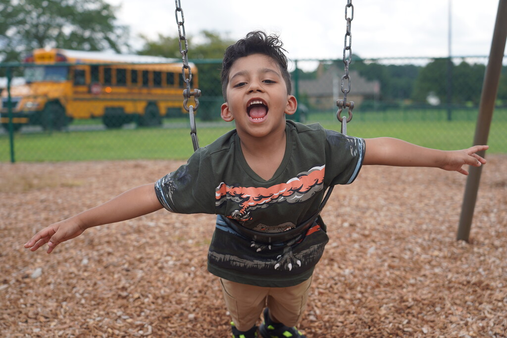 Child with arms wide open on a swing, mouth open. Behind them is a school bus.