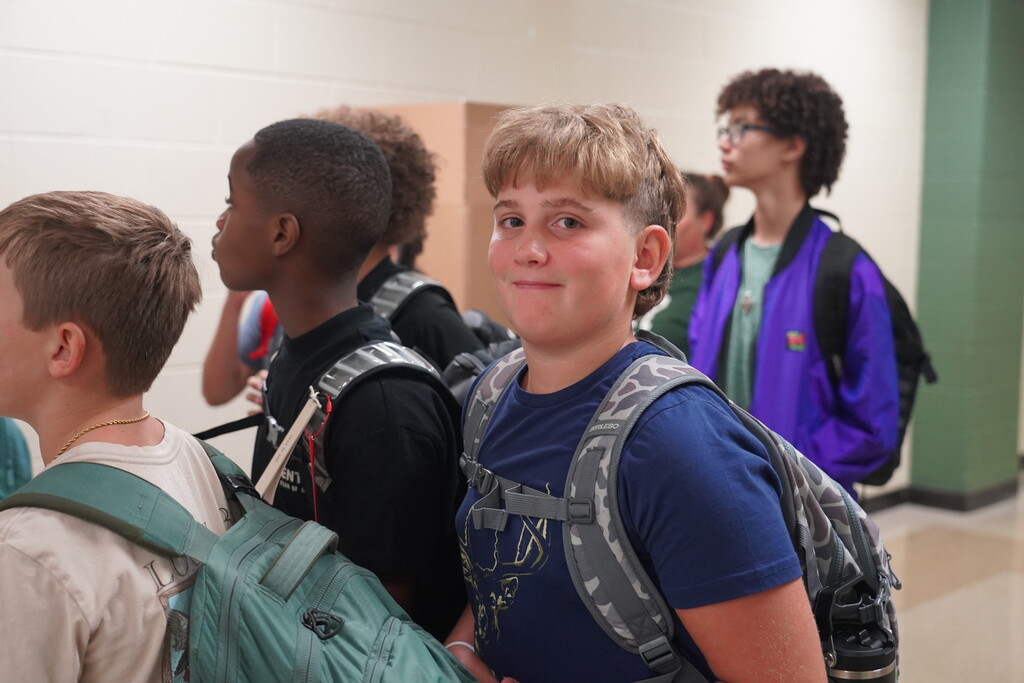 Several children, each with a backpack, stand in a hallway. Some are looking away while others are smiling.