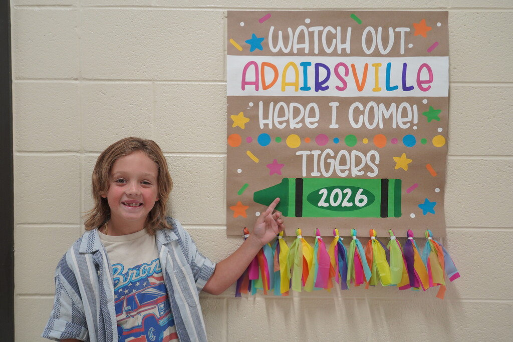 A young child smiles and points at a colorful poster featuring a crayon and the text "Watch out Adairsville, here I come! Tigers 2026."