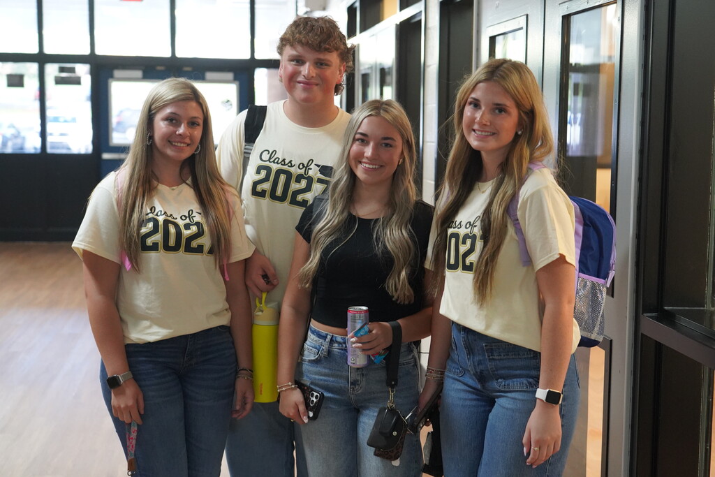 Four women in jeans pose for a photo. One wears a "2021" shirt, another a "2022" shirt. One holds a drink and handbag.