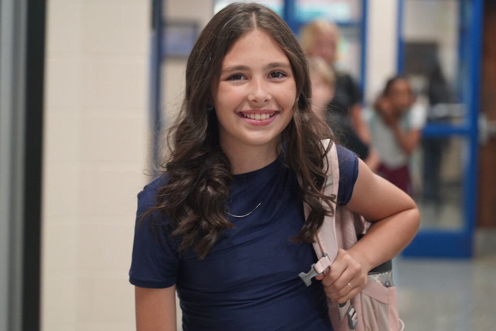 A girl with curly hair in a navy blue shirt, smiling, stands in a hallway with others.