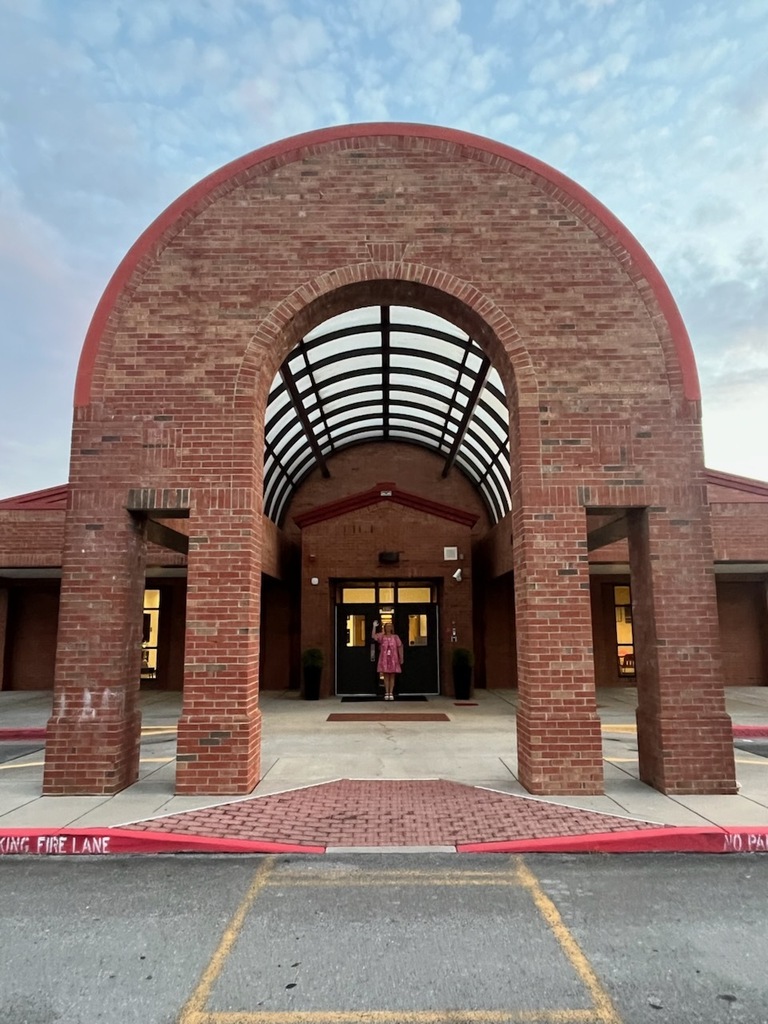 Brick archway with a glass roof in front of a building, with a teacher waving from the front door. A bright blue sky in the background shows that it is daytime and pleasant weather.