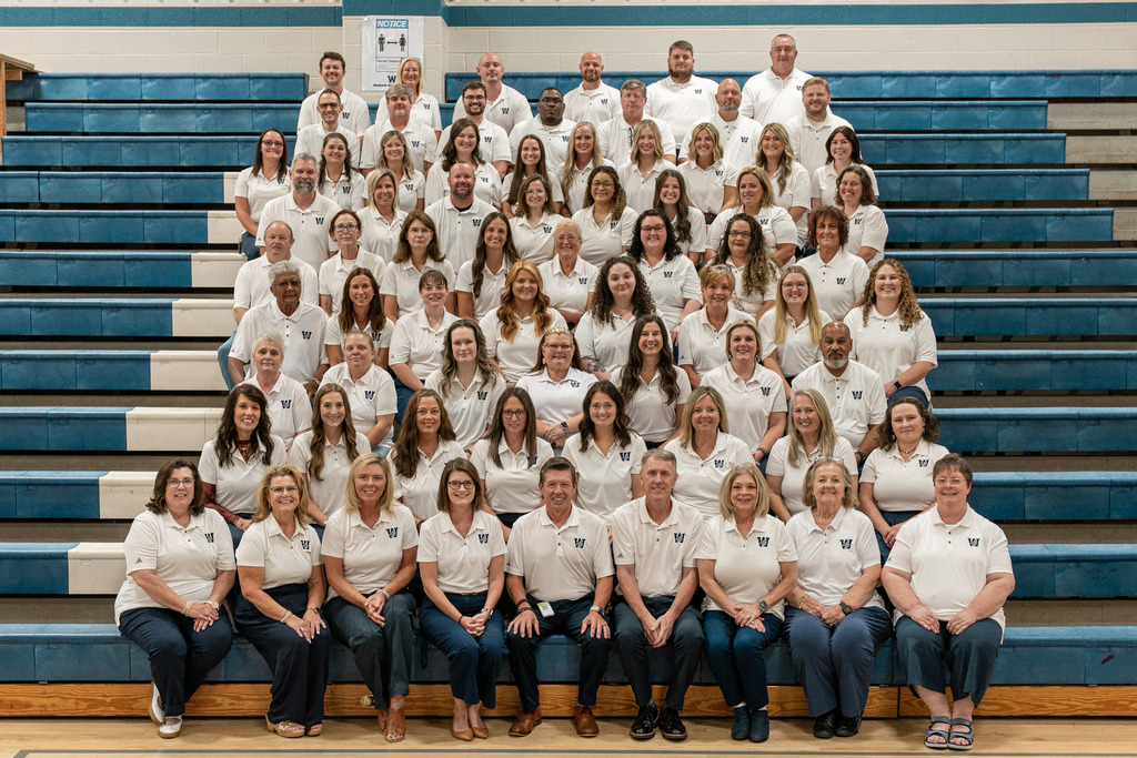 A group of people in matching white shirts and jeans pose for a photo on bleachers.
