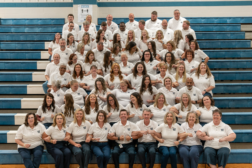 A large group of people in white shirts and jeans pose for a photo on bleachers.